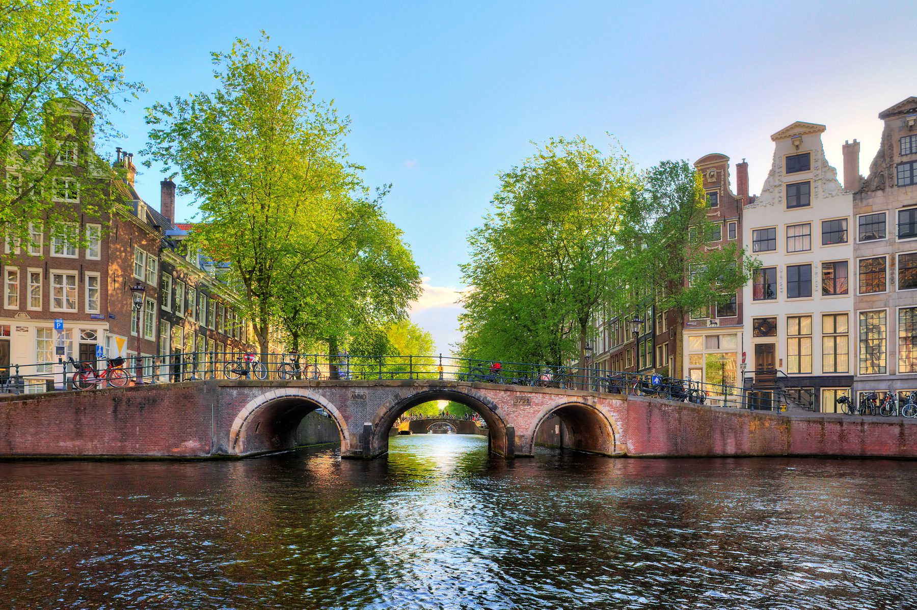 Een stenen brug met drie bogen over een Amsterdamse gracht, met grachtenpanden en bomen langs de kade